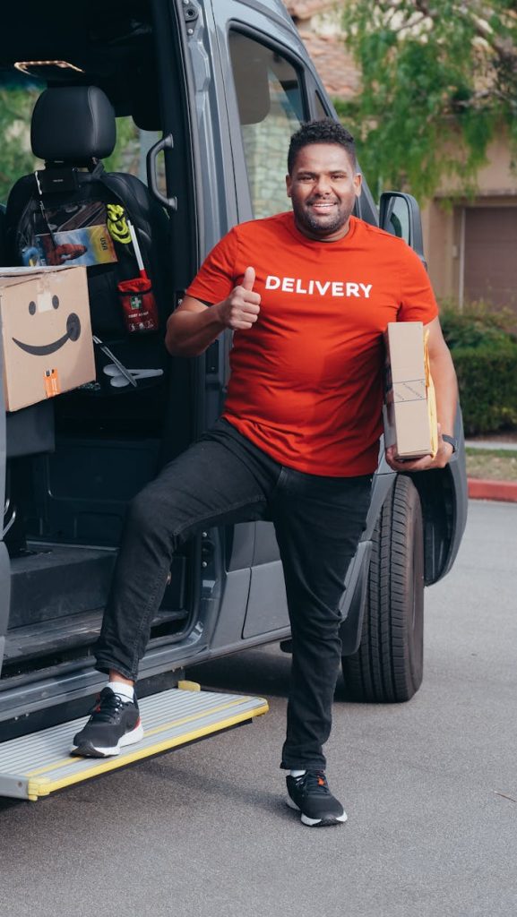 Cheerful delivery man in a red shirt holding a package and giving a thumbs up by his van.